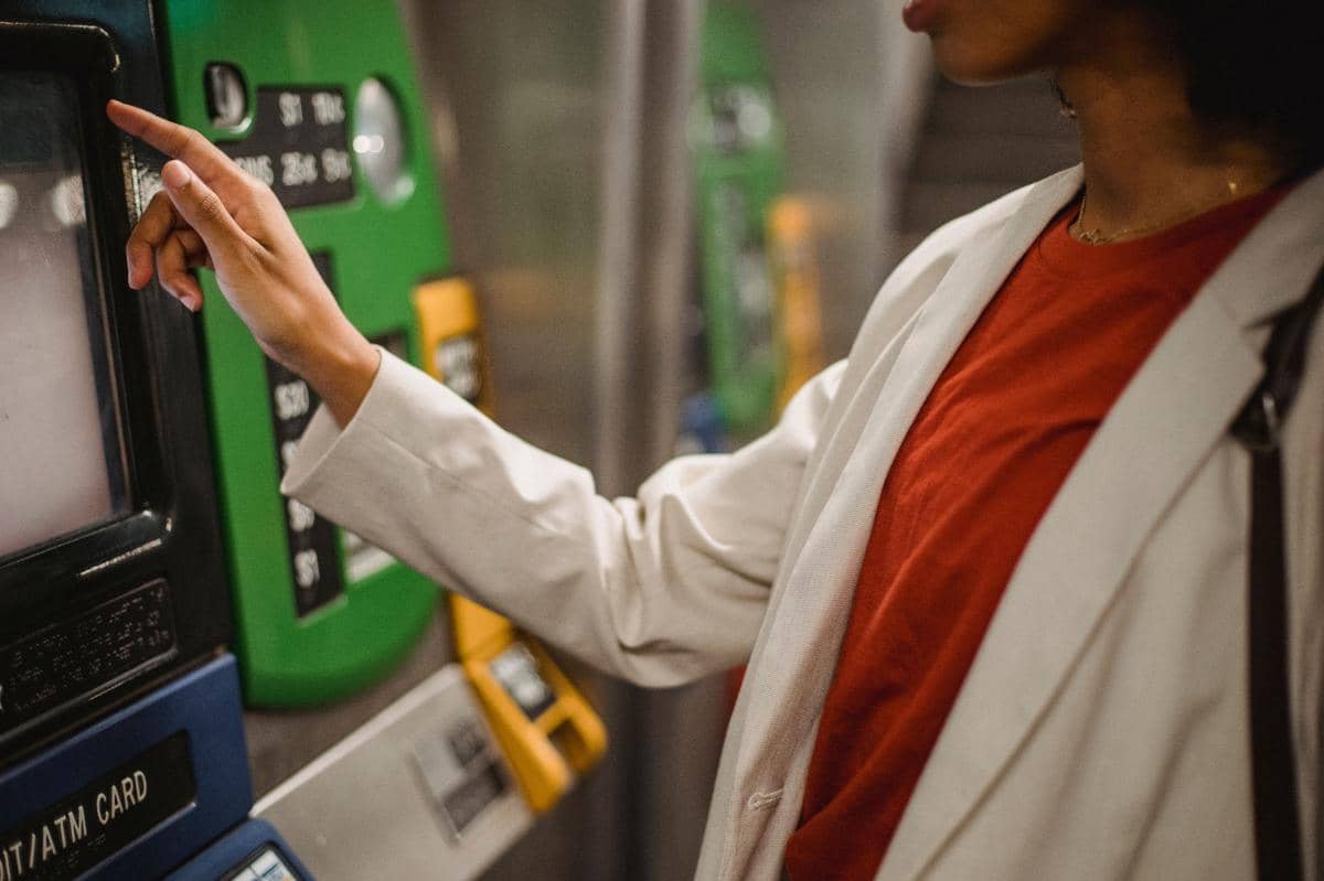 Seseorang mengenakan jas krem dan kaus merah sedang menyentuh layar mesin ATM berwarna hijau di area publik.