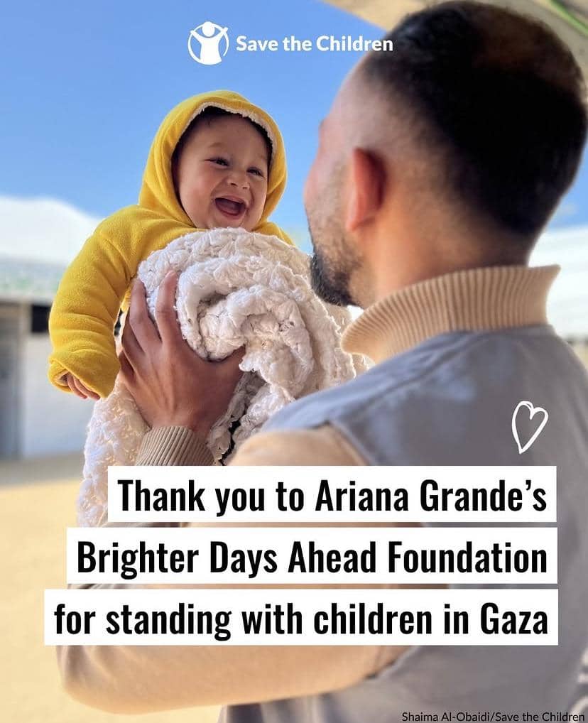 Seorang pria mengangkat bayi yang tersenyum mengenakan jaket kuning di bawah langit cerah dengan logo Save the Children di atasnya.