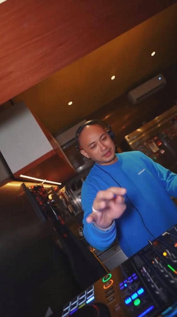 Seorang DJ mengenakan sweater biru sedang memainkan musik di meja kontrol DJ dalam suasana pesta perpisahan Indonesian Idol Season 14.