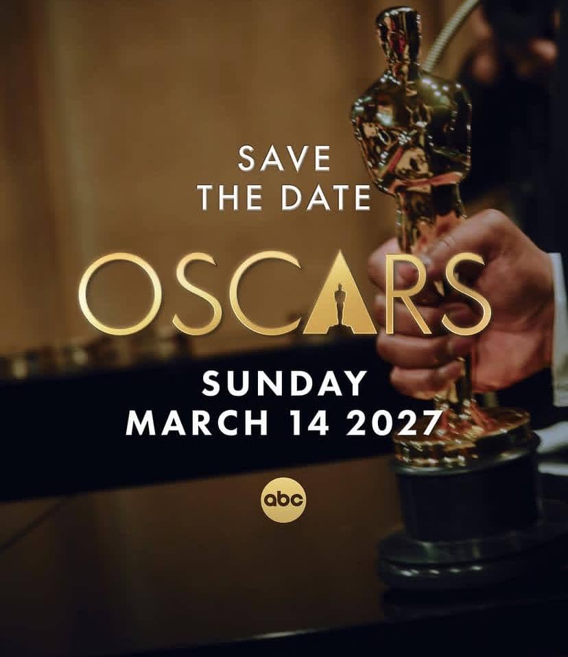 Tangan seseorang memegang piala Oscar berwarna emas dengan tulisan pengumuman tanggal acara Oscars pada 14 Maret 2027.