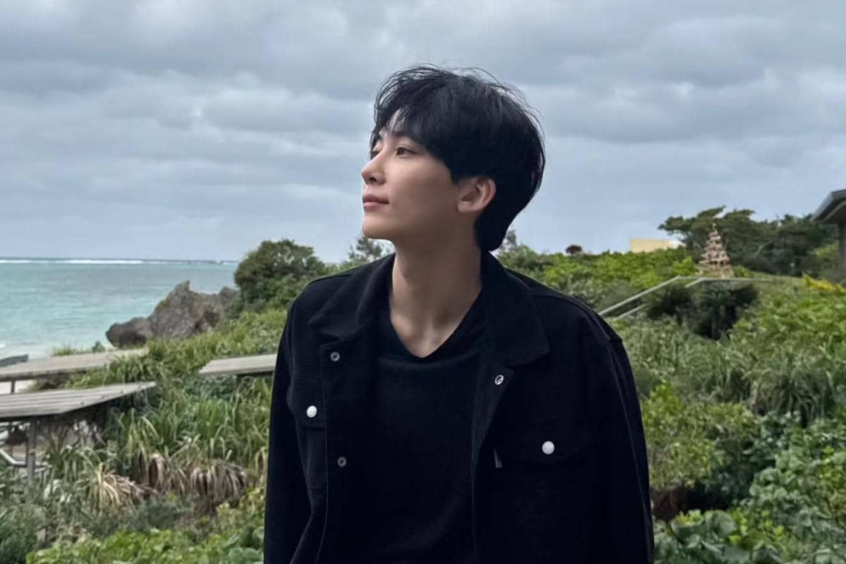 Seorang pria berambut hitam mengenakan jaket hitam berdiri di tepi pantai dengan latar pepohonan hijau dan langit berawan.