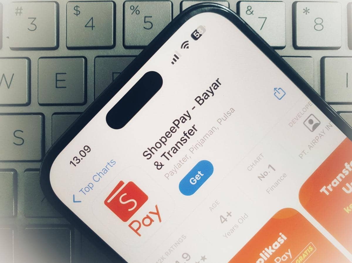 Tampilan aplikasi ShopeePay di layar ponsel yang diletakkan di atas keyboard laptop, menampilkan opsi unduh di App Store.