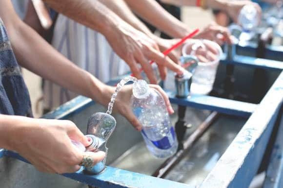 Dekat tampak samping tangan beberapa orang sedang memegang botol plastik transparan untuk diisi ulang dengan air bersih yang mengalir dari dispenser fasilitas umum.
