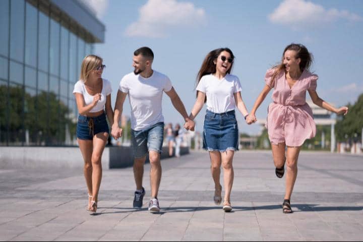 Empat orang remaja laki-laki dan perempuan berpakaian kasual sedang bergandengan tangan sambil berlari dan tertawa bersama di area pejalan kaki di depan gedung kaca modern.