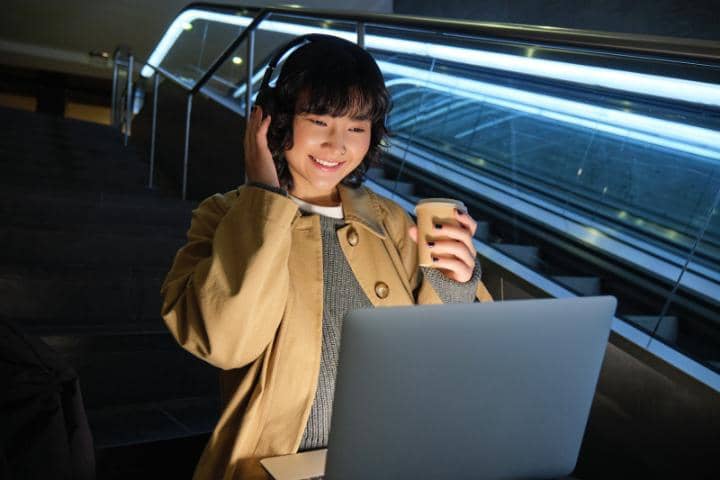 Seorang remaja perempuan menggunakan headphone sedang tersenyum melihat layar laptop sambil memegang segelas kopi di tangga.