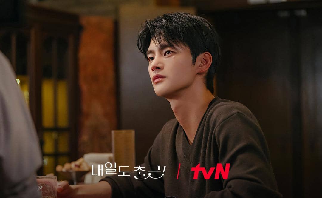 Seorang pria duduk di meja restoran dengan ekspresi serius, mengenakan sweater cokelat dalam adegan drama televisi tvN.