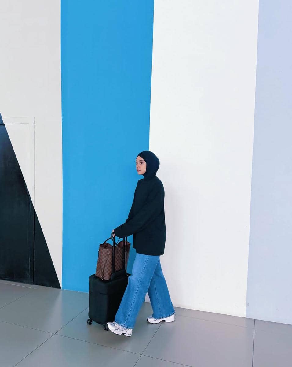 Seorang wanita berhijab mengenakan sweater hitam dan celana jeans longgar sedang menarik koper kecil di dekat dinding berwarna biru dan putih.