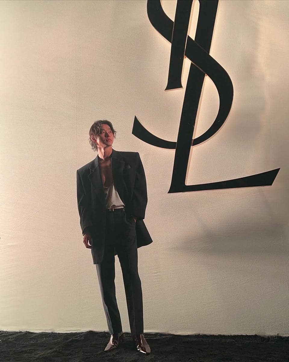 Seorang pria mengenakan jas hitam modern dengan kemeja putih terbuka di depan logo besar YSL di latar belakang berwarna krem.