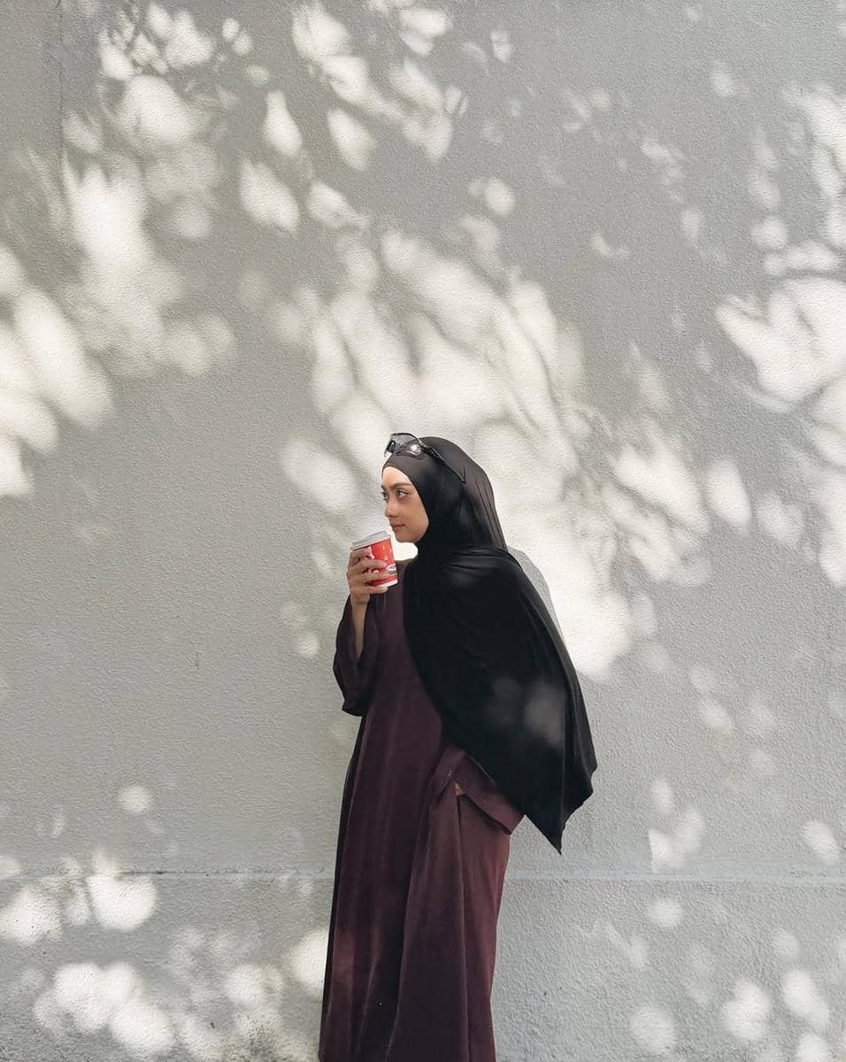 Seorang wanita berhijab hitam mengenakan dress panjang berwarna cokelat sambil memegang gelas minuman di bawah bayangan pepohonan.