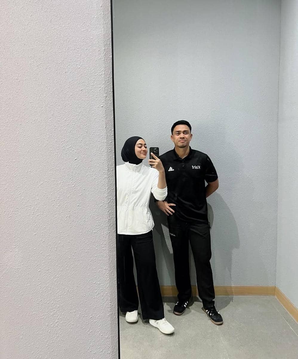 Seorang wanita berhijab bergaya minimalis mengenakan jaket putih dan celana hitam berpose di cermin bersama pria berpakaian hitam.