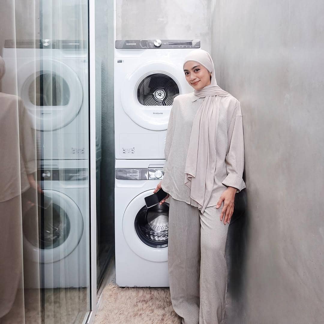 Seorang wanita berhijab bergaya minimalis dengan busana berwarna netral berdiri di samping mesin cuci tumpuk di ruangan modern.