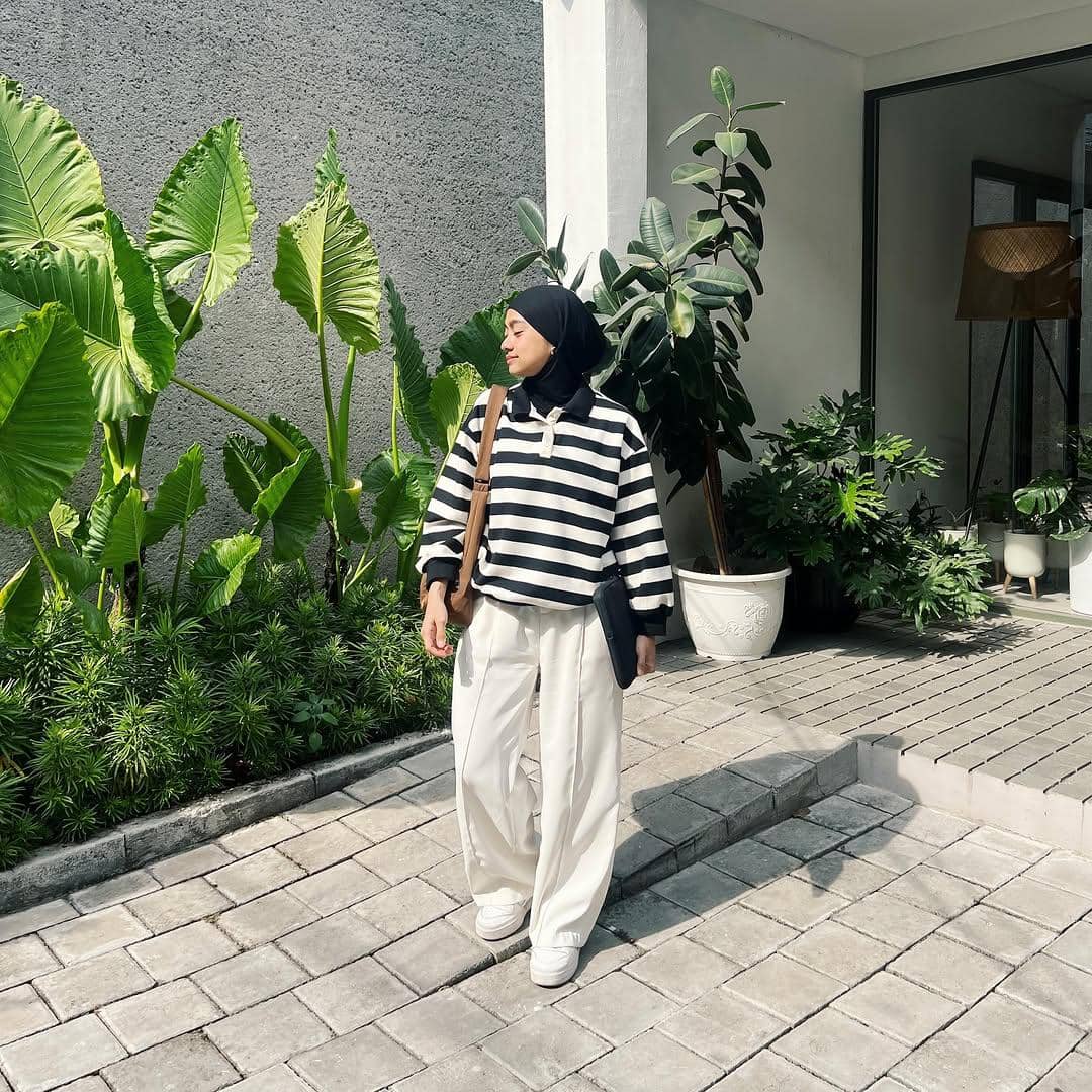 Perempuan berhijab mengenakan atasan garis hitam putih dan celana panjang putih, berdiri di halaman dengan tanaman hijau di sekitarnya.