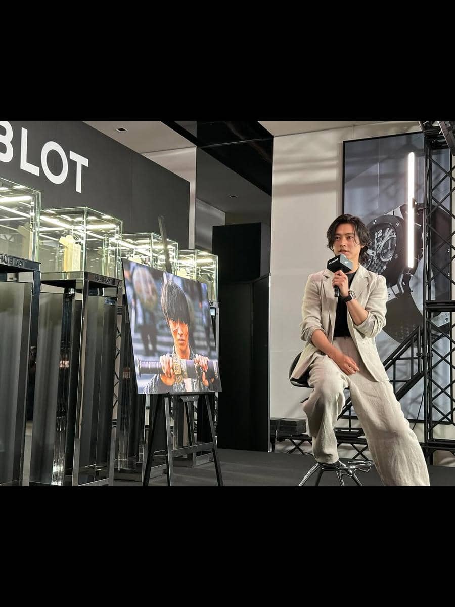 Seorang pria mengenakan jas modern berwarna terang duduk di kursi sambil memegang mikrofon di acara dengan latar merek Hublot.