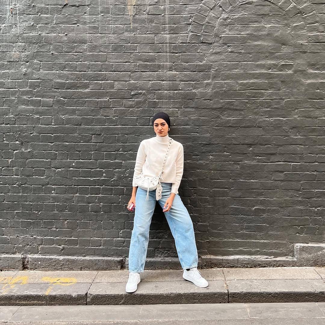 Perempuan berhijab mengenakan sweater putih, celana jeans longgar, dan sepatu putih berpose di depan dinding bata abu-abu.