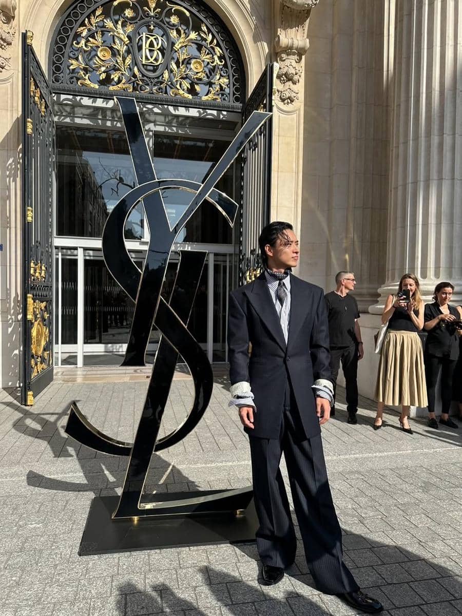 Seorang pria mengenakan jas modern berwarna gelap berdiri di depan instalasi logo YSL besar di luar gedung bergaya klasik dengan gerbang besi berornamen emas.
