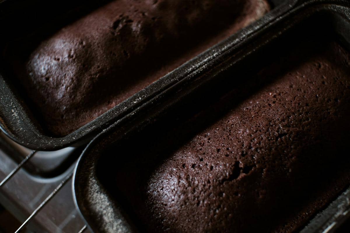 Dua loyang berisi roti zukini cokelat yang baru dipanggang dengan permukaan lembap dan bertekstur halus berwarna cokelat pekat.