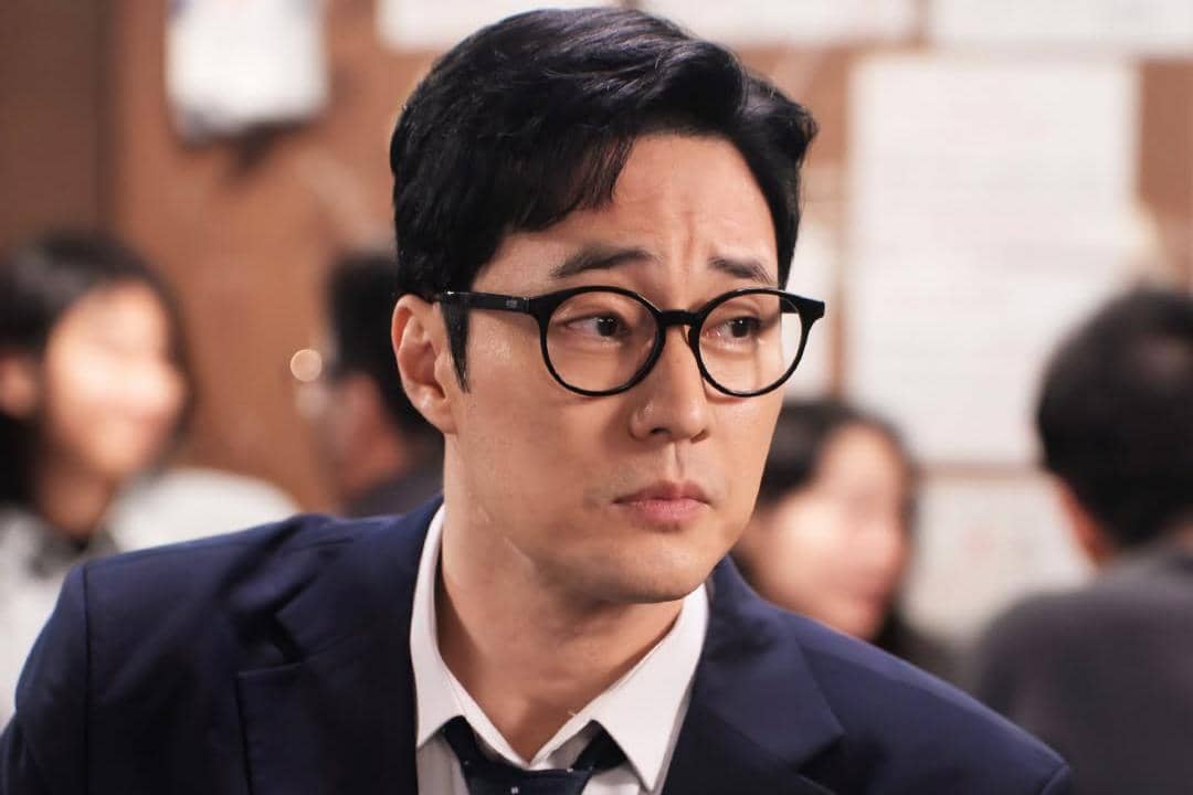 Seorang pria berkacamata mengenakan setelan jas biru dan dasi hitam terlihat serius dalam adegan drama Agent Kim Reactivated.