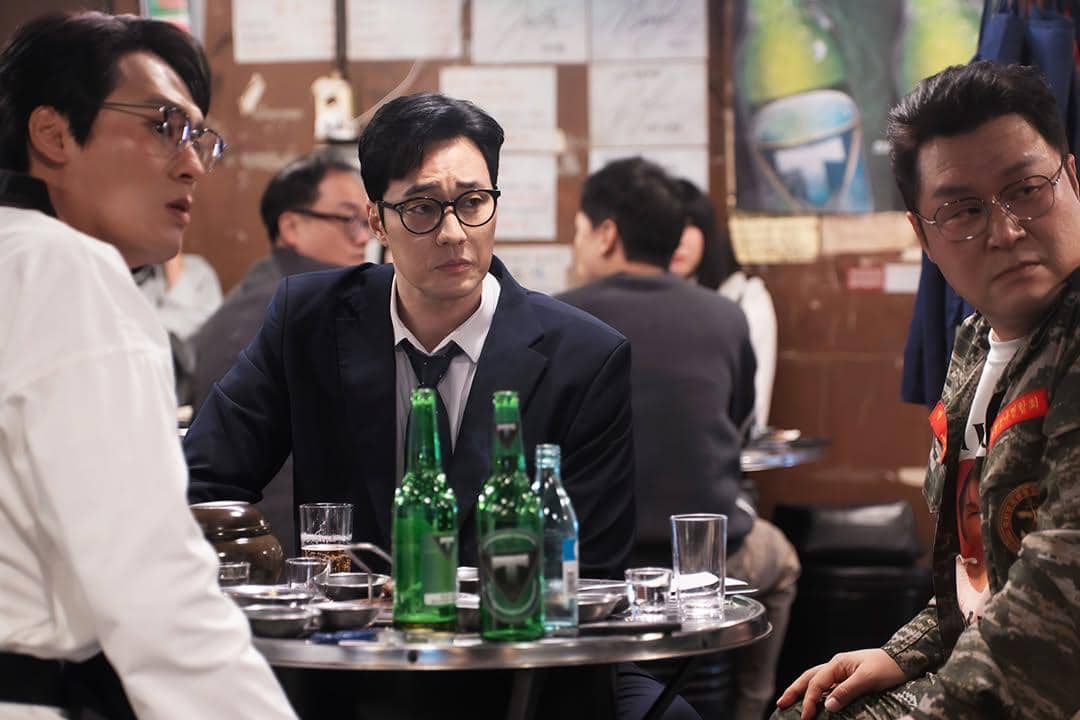 Tiga pria duduk di meja restoran kecil dengan botol minuman di atas meja dalam adegan drama Agent Kim Reactivated.