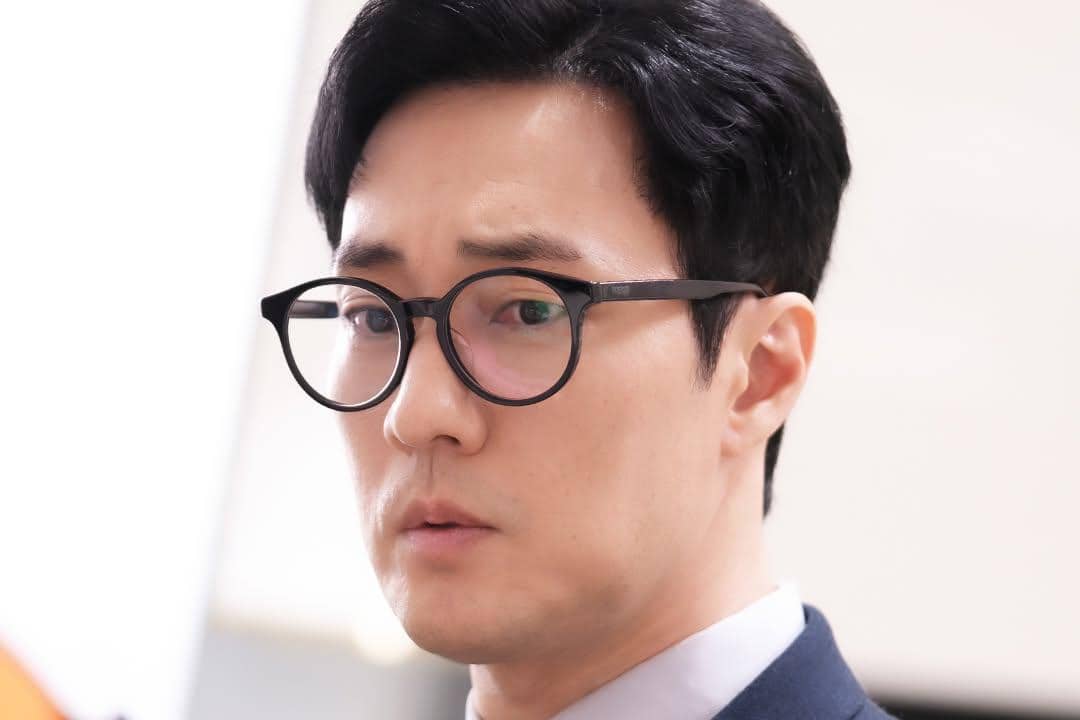 Seorang pria berpenampilan rapi mengenakan kacamata bulat hitam dan setelan jas, tampak serius dalam adegan drama Agent Kim Reactivated.