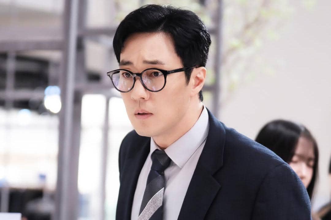 Seorang pria mengenakan setelan jas dan kacamata berdiri di dalam ruangan kantor dengan ekspresi serius dalam adegan drama Agent Kim Reactivated.