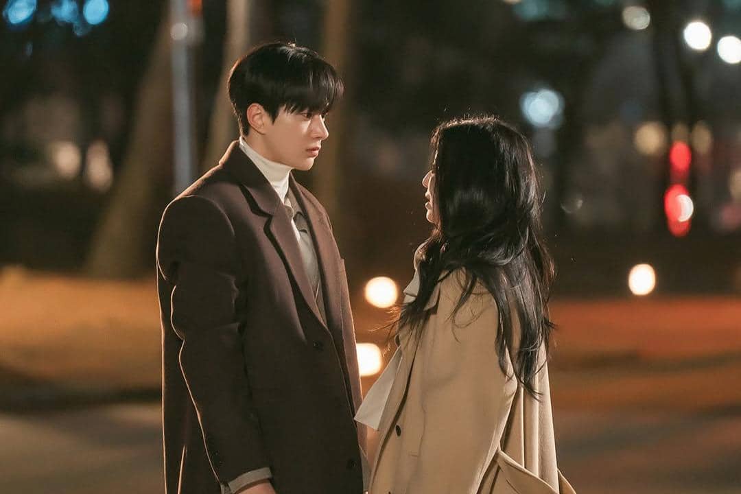 Dua pemeran drama Love In Sync berdiri saling berhadapan di malam hari dengan latar lampu kota yang hangat dan suasana romantis.