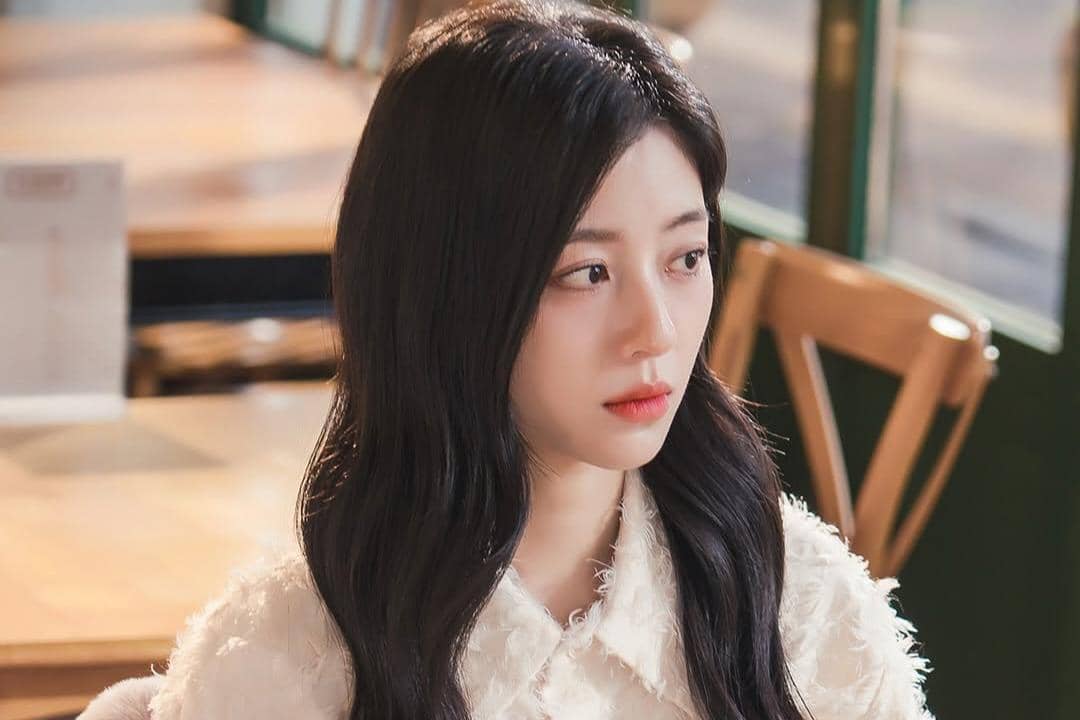 Seorang wanita berambut panjang hitam mengenakan blus putih duduk di kafe dengan pencahayaan hangat, menatap ke samping dengan ekspresi tenang.