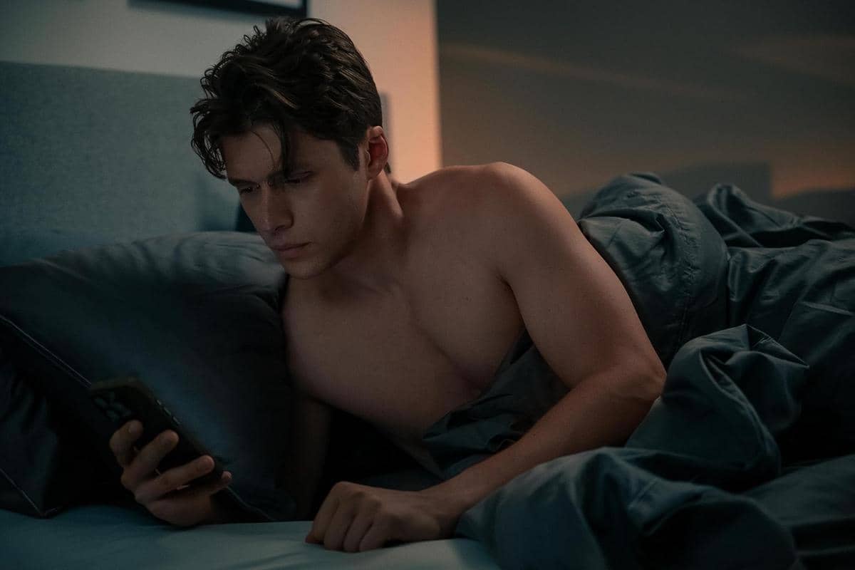 Seorang pria tanpa baju berbaring di tempat tidur sambil menatap ponsel dengan ekspresi serius dalam adegan film Voicemails for Isabelle.