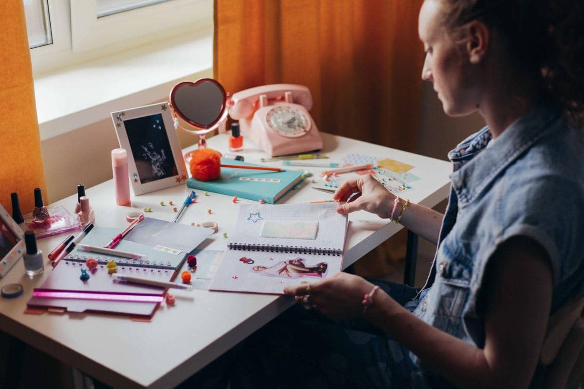 Seorang perempuan duduk di meja dekat jendela sedang membuat scrapbook dengan berbagai alat tulis, kertas warna, dan hiasan kecil.