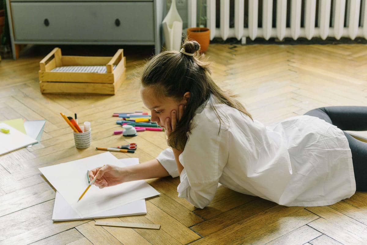 Seorang perempuan berbaring di lantai kayu sambil menggambar di atas kertas dengan pensil, dikelilingi alat tulis warna-warni.