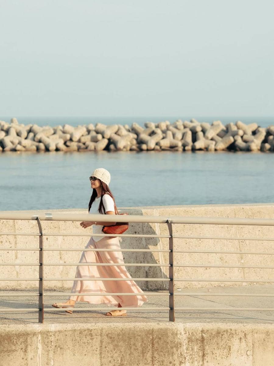 Seorang wanita berjalan di tepi pantai Busan dengan mengenakan rok panjang berwarna lembut, topi rajut, dan tas cokelat di bawah sinar matahari.