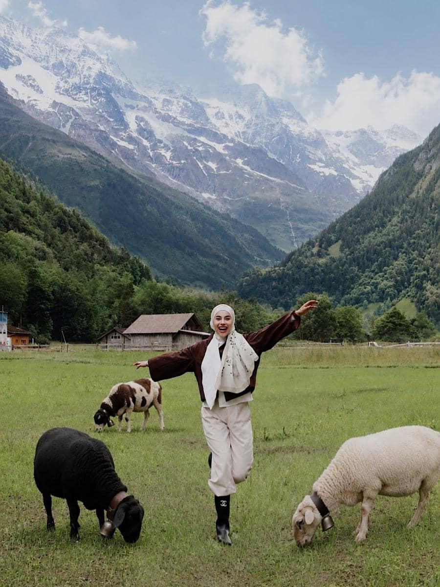Seorang perempuan berhijab berpose ceria di padang rumput hijau dengan domba dan sapi di Desa Lauterbrunnen berlatar pegunungan bersalju.