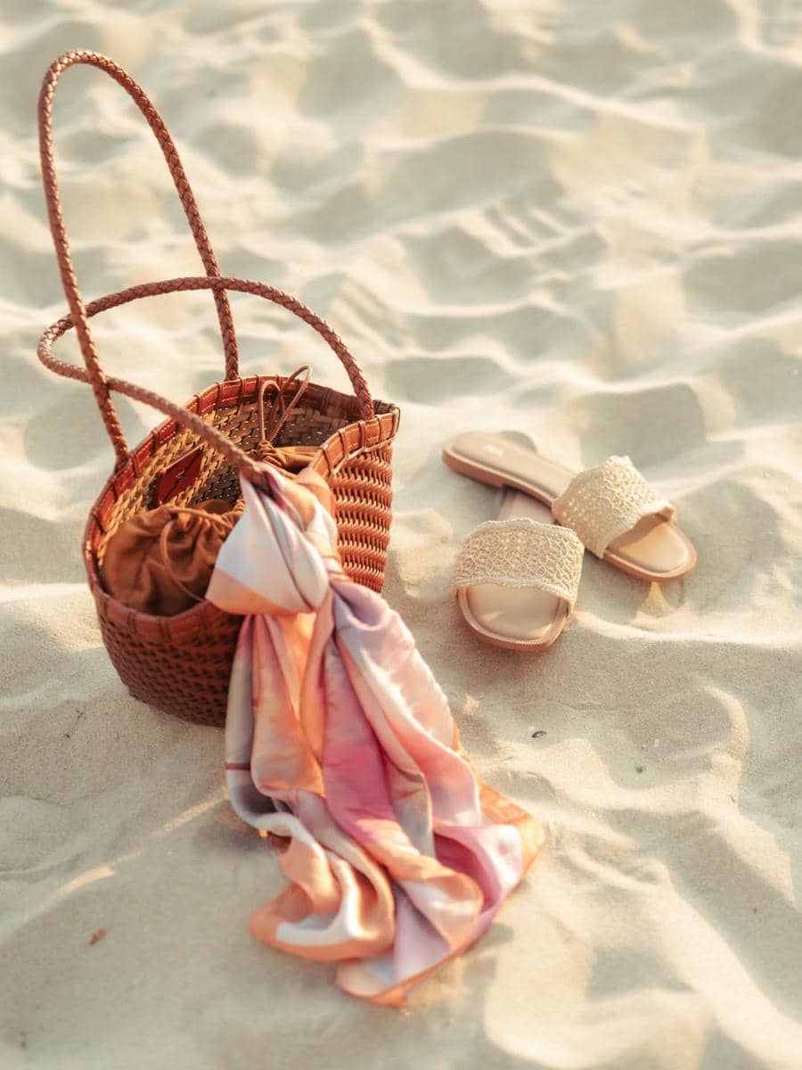 Tas rotan dengan selendang warna pastel dan sepasang sandal anyaman tergeletak di pasir pantai saat matahari sore.