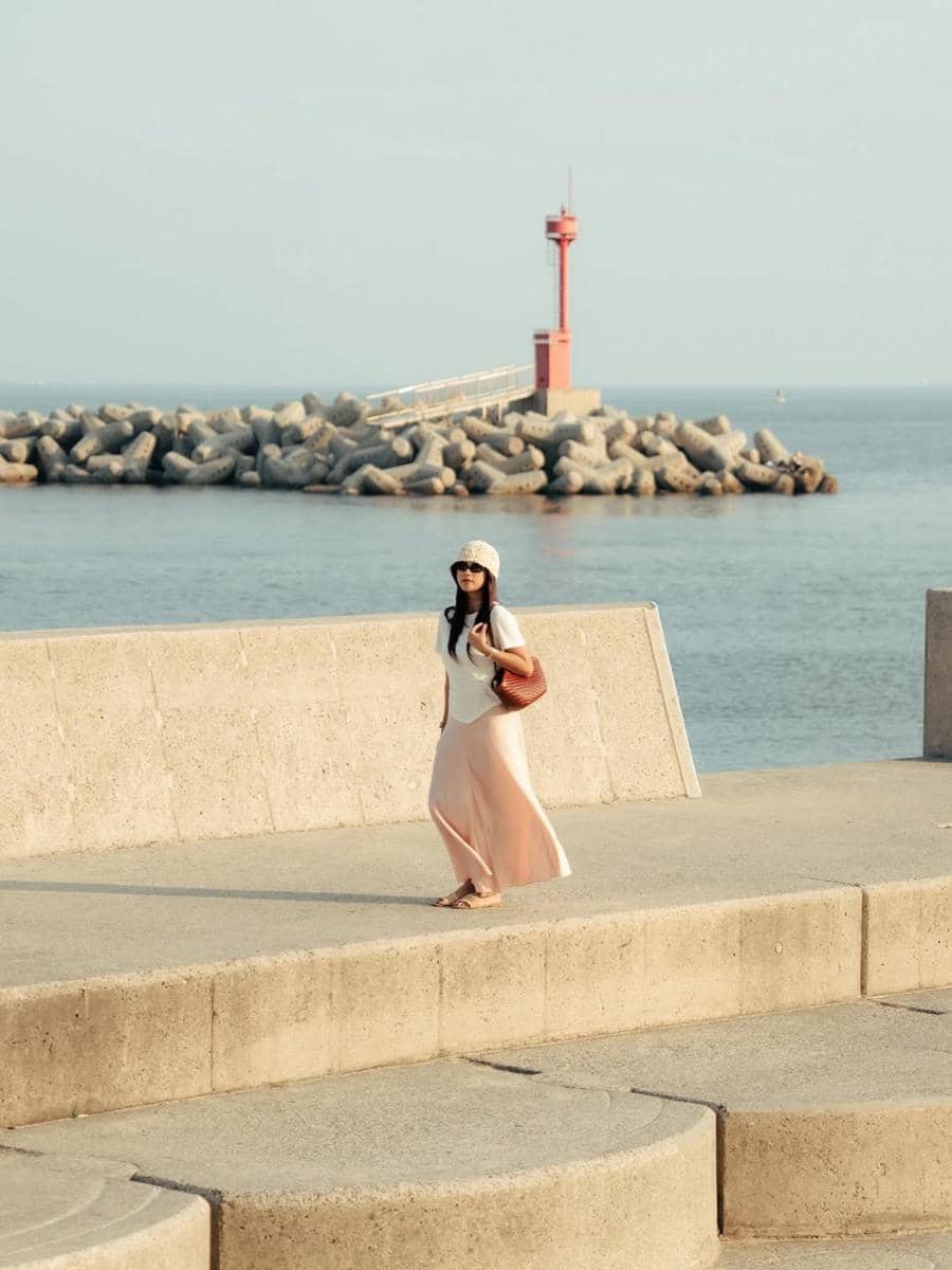 Seorang perempuan berjalan di tepi pantai Busan dengan latar belakang mercusuar merah dan pemecah ombak di bawah langit cerah.