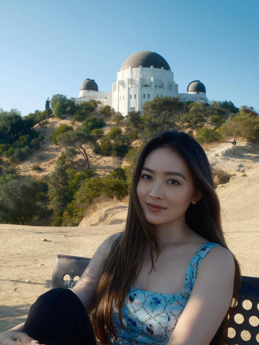 Seorang perempuan berpose di depan Griffith Observatory di Los Angeles dengan latar langit cerah dan pepohonan di sekitarnya.