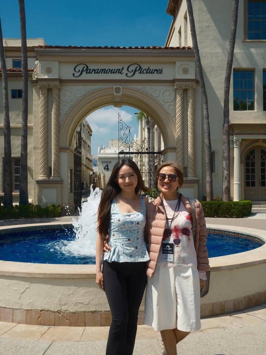 Dua wanita berpose di depan gerbang Paramount Pictures dengan air mancur dan bangunan bergaya klasik di Hollywood.