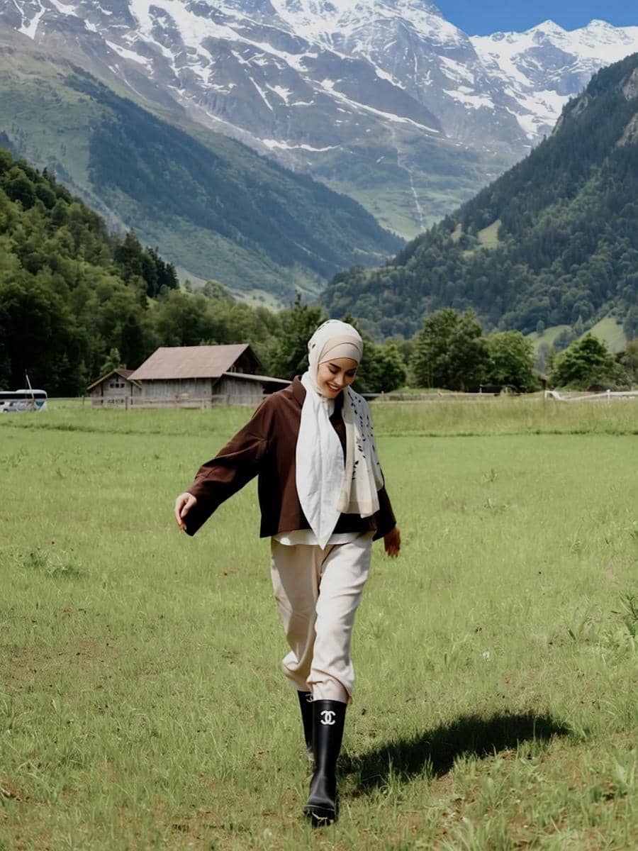Seorang perempuan berhijab berjalan di padang rumput hijau dengan latar pegunungan bersalju di Desa Lauterbrunnen, Swiss.