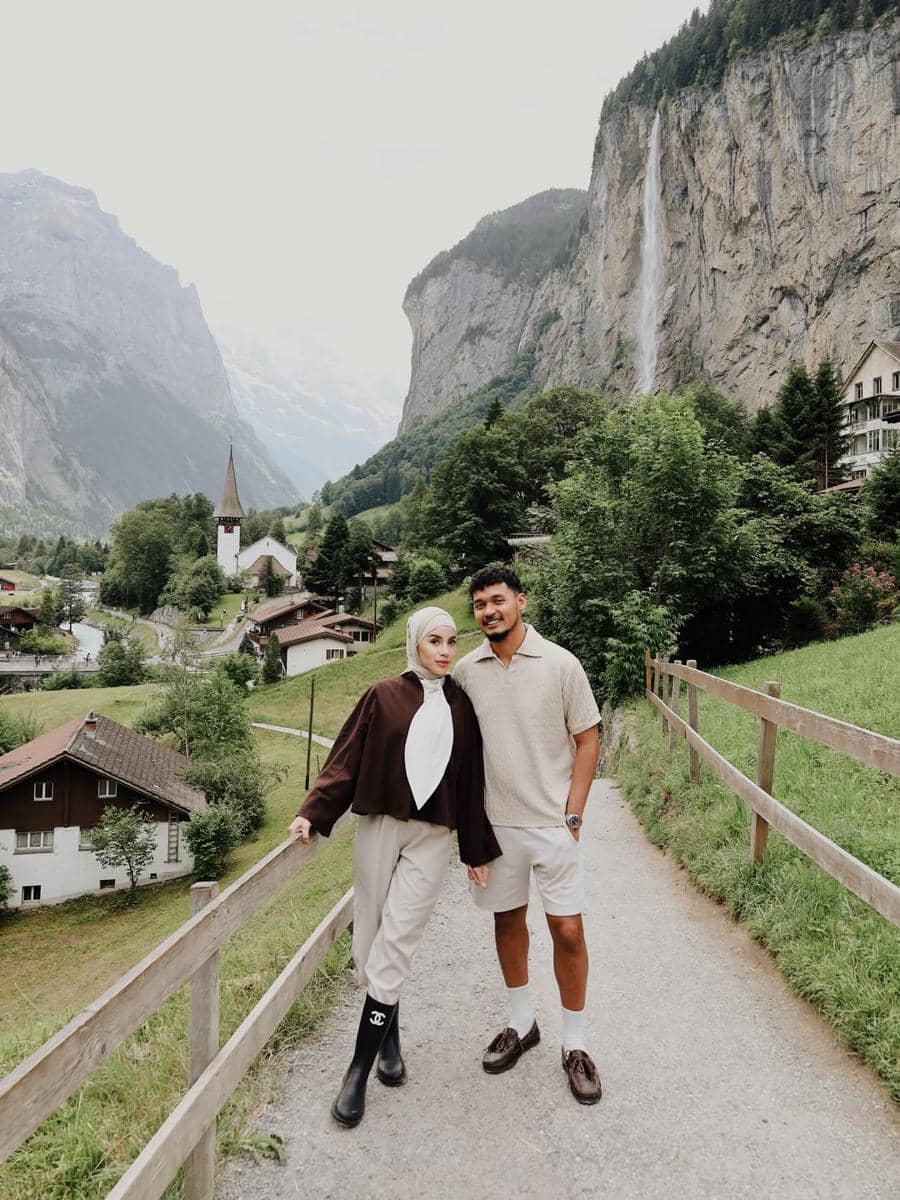 Sepasang wisatawan berpose di jalan pedesaan Lauterbrunnen dengan latar tebing tinggi, air terjun, dan rumah khas Swiss di sekitarnya.