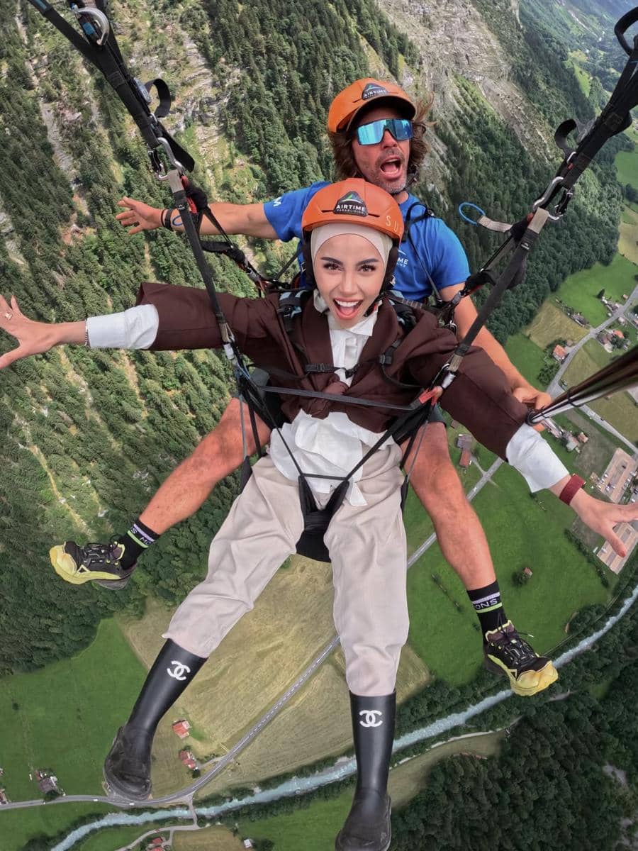 Seorang wanita berhijab dan instruktur paragliding terbang di atas lembah hijau Desa Lauterbrunnen dengan ekspresi gembira.