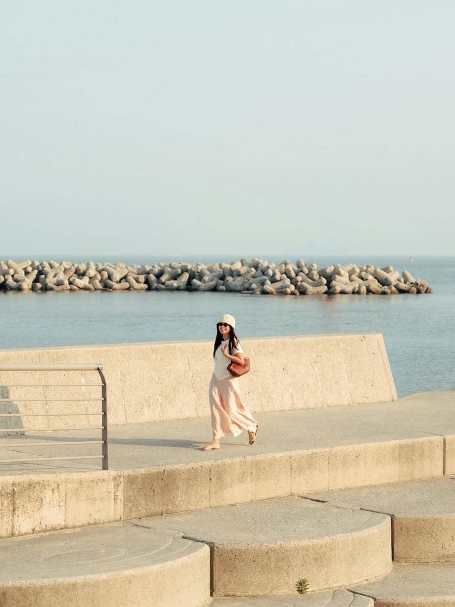 Seorang wanita berjalan santai di tepi pantai Busan dengan latar laut tenang dan pemecah ombak beton di bawah langit cerah.