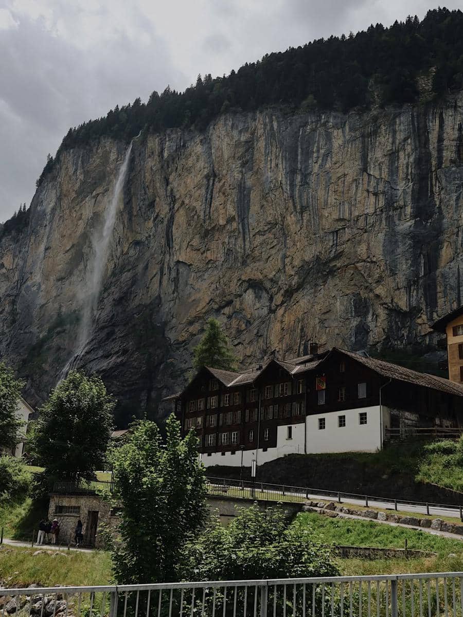 Pemandangan indah Desa Lauterbrunnen dengan tebing tinggi, air terjun mengalir, dan rumah kayu tradisional di bawah langit mendung.