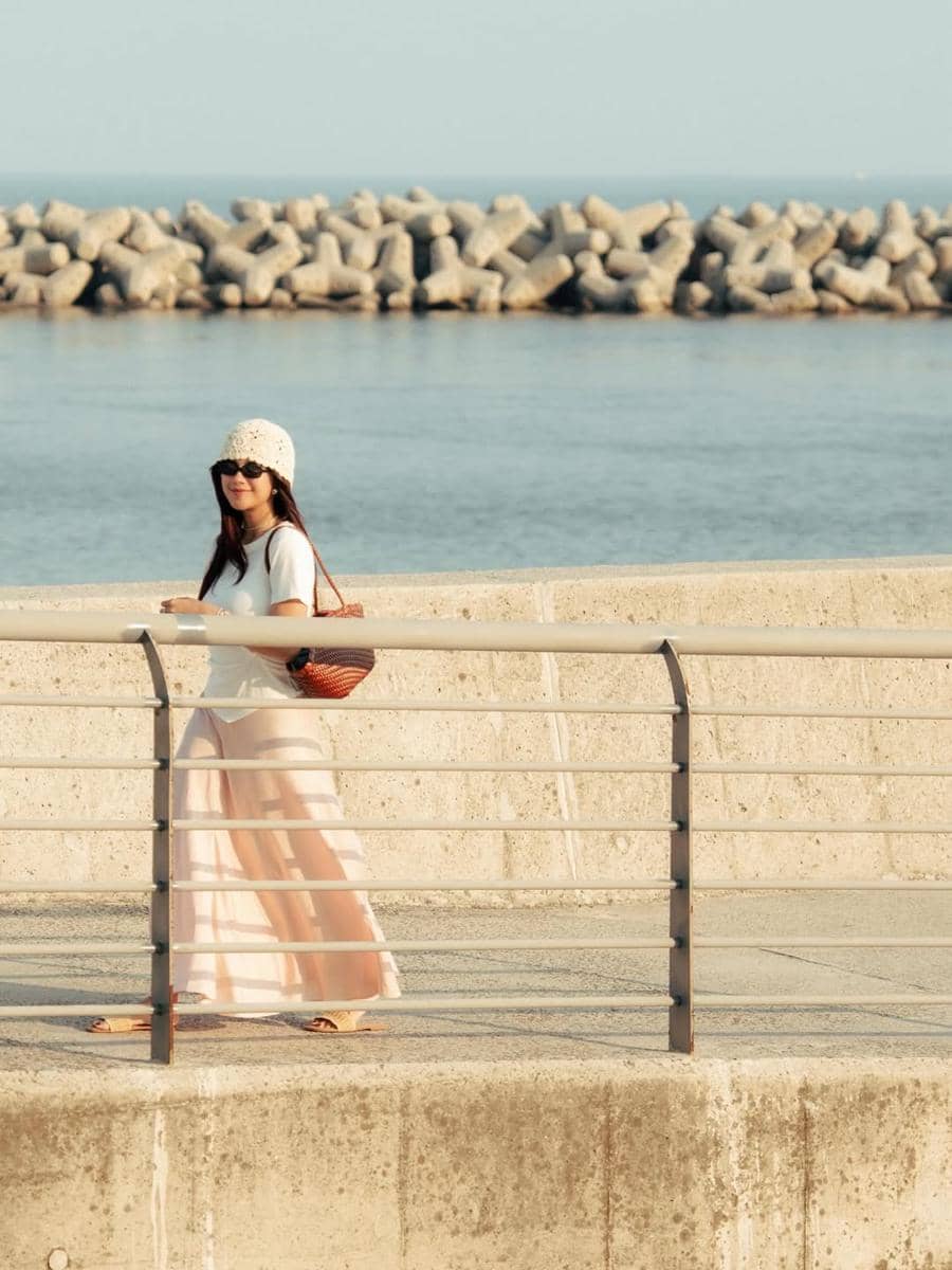 Seorang wanita mengenakan topi rajut dan rok panjang berjalan di tepi pantai dengan latar laut dan pemecah ombak di Busan.