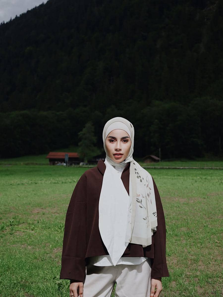 Seorang perempuan berhijab berpose di tengah padang rumput hijau dengan latar pegunungan dan pepohonan di Desa Lauterbrunnen.