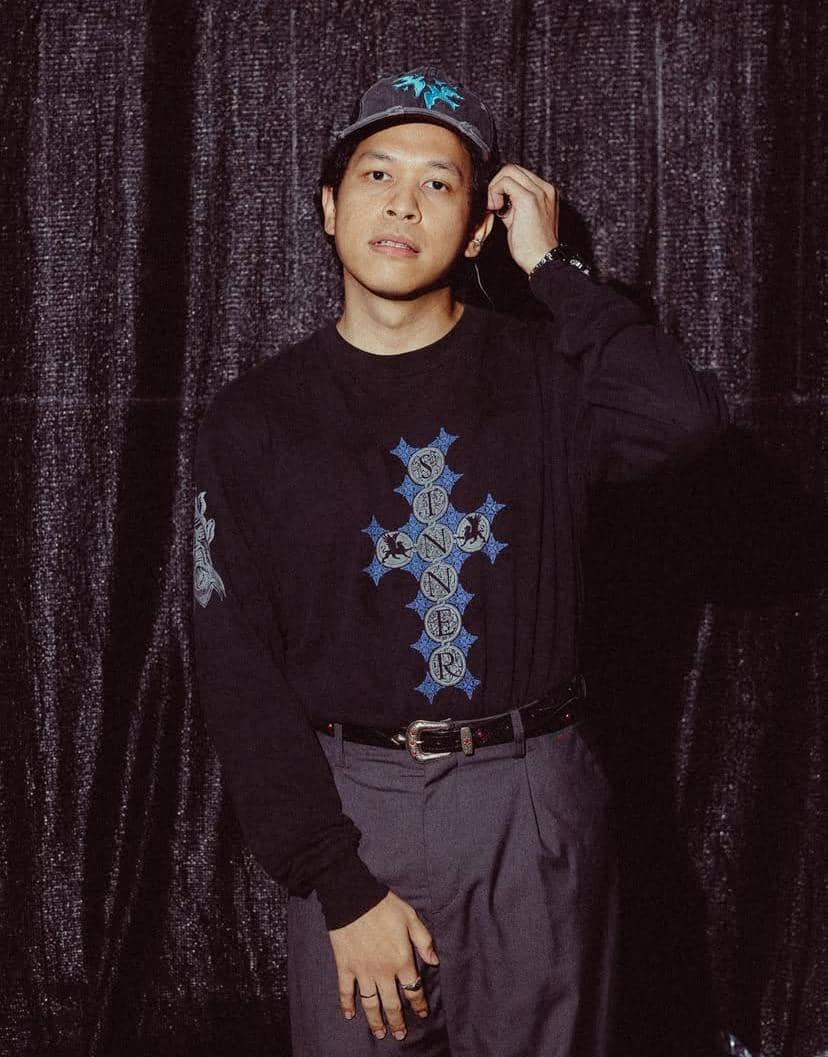 Seorang pria mengenakan topi abu-abu dan kaus hitam bergambar salib biru dengan tulisan, berdiri di depan tirai hitam.