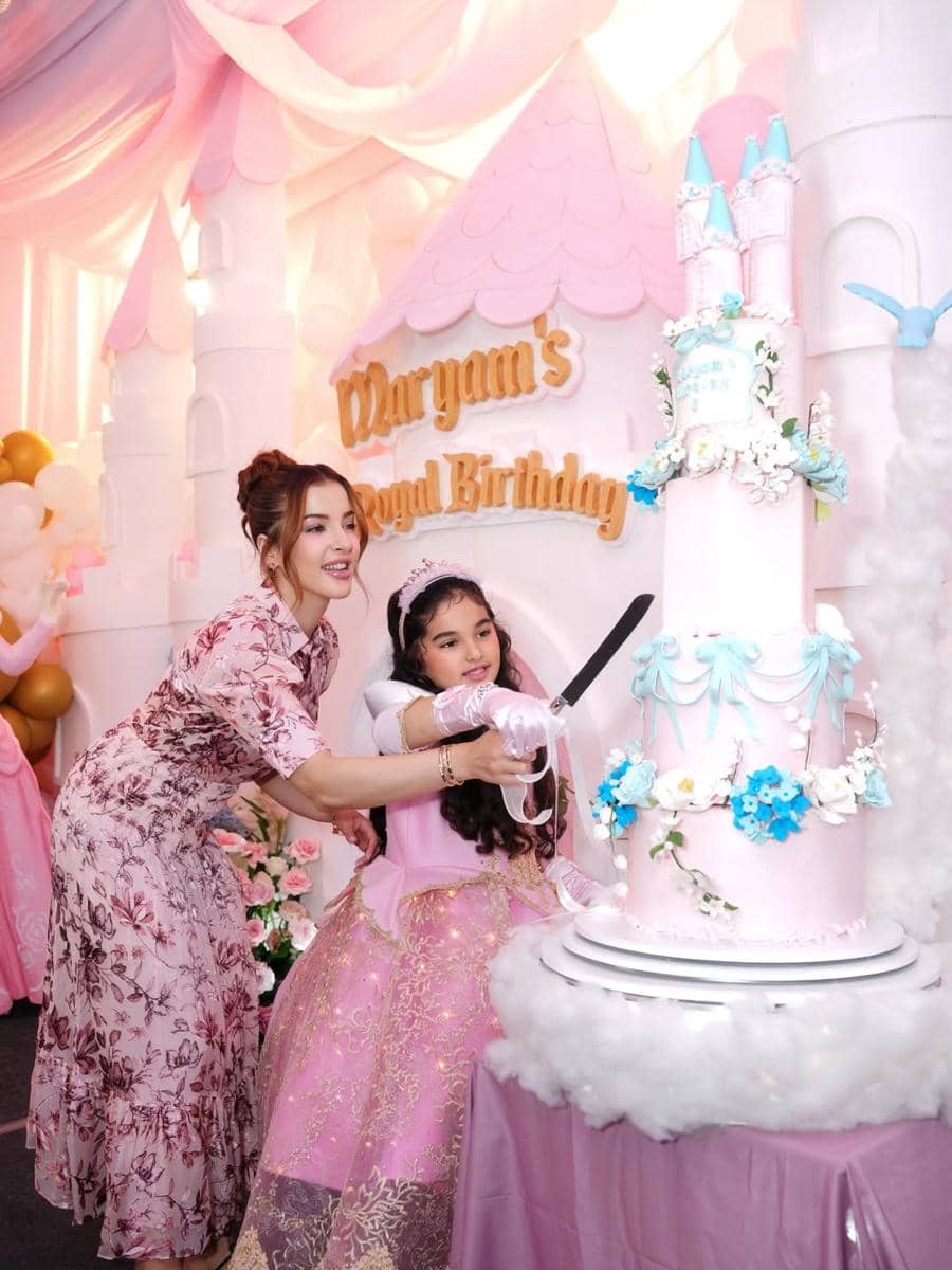 Seorang wanita dan anak perempuan mengenakan gaun berwarna pink sedang memotong kue ulang tahun tinggi bertema kastel di pesta ulang tahun keenam.