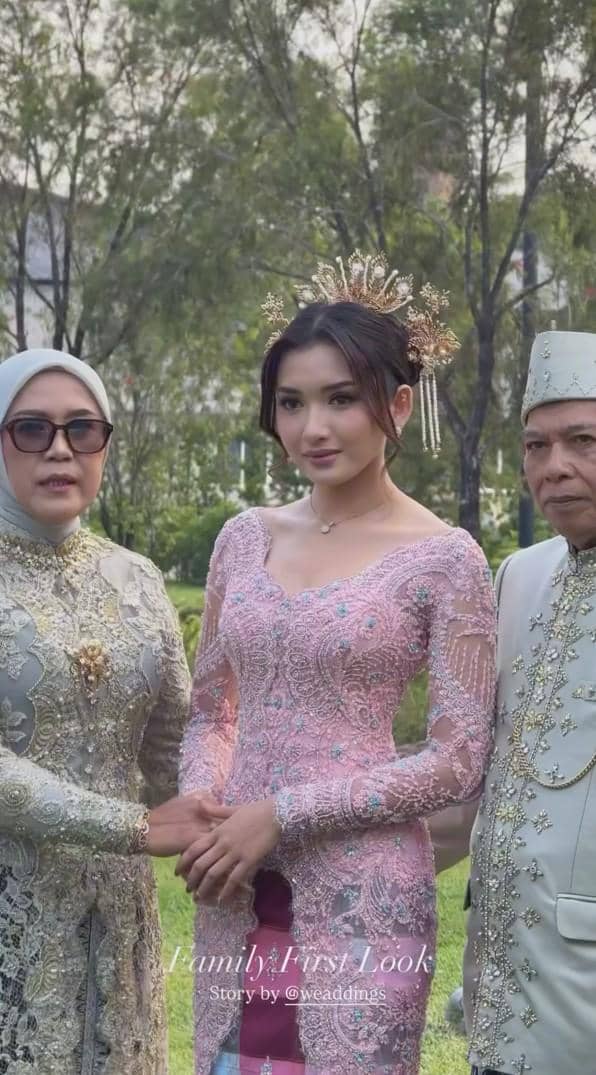 Seorang perempuan mengenakan kebaya pink berhias mahkota emas berpose bersama dua orang lanjut usia dalam balutan busana tradisional.