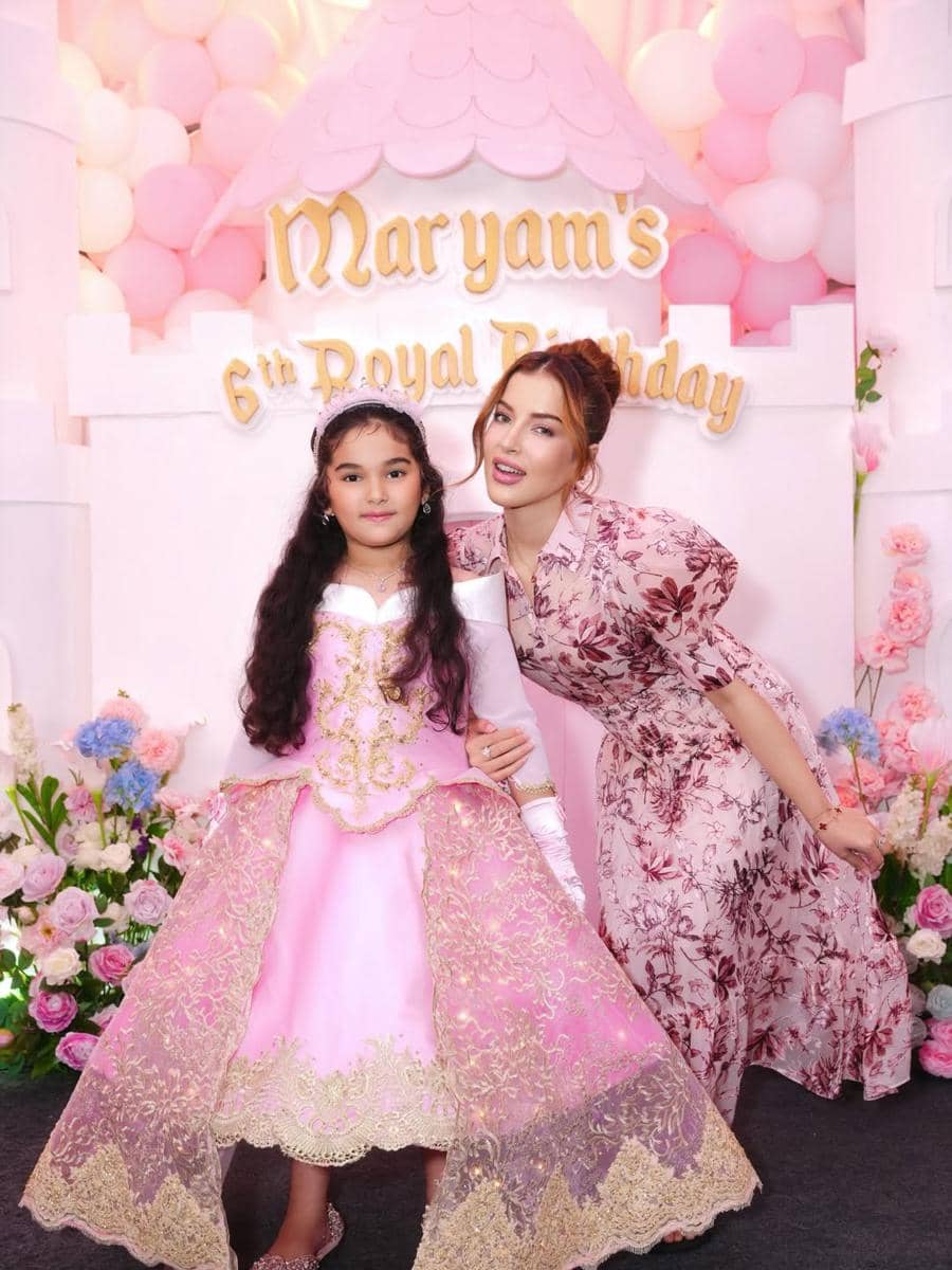 Seorang anak perempuan mengenakan gaun pink bertema putri berdiri bersama seorang wanita di pesta ulang tahun ke-6 dengan dekorasi balon dan bunga.
