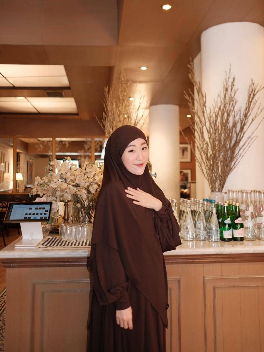 Seorang wanita berhijab cokelat berdiri di depan meja bar dengan botol kaca dan dekorasi bunga kering di dalam ruangan bernuansa hangat.