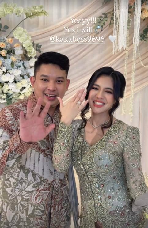 Sepasang pria dan wanita mengenakan busana tradisional berpose sambil menunjukkan cincin lamaran di acara pertunangan mereka.