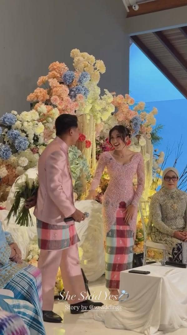 Pasangan mengenakan busana tradisional warna senada bernuansa merah muda saat acara lamaran dengan latar bunga warna-warni.