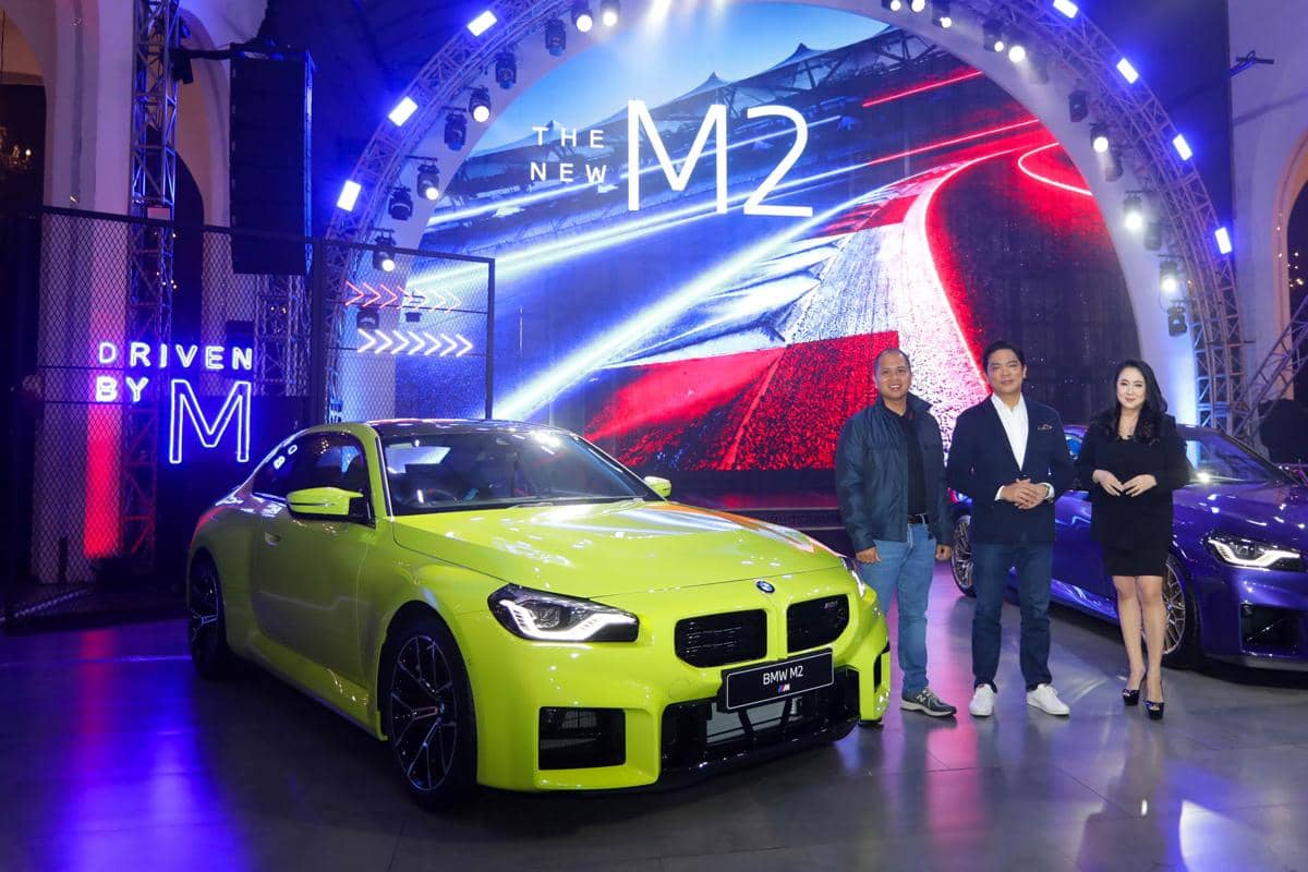 BMW M2 terbaru berwarna kuning dipamerkan di acara peluncuran dengan latar panggung bertema balap dan pencahayaan biru merah.
